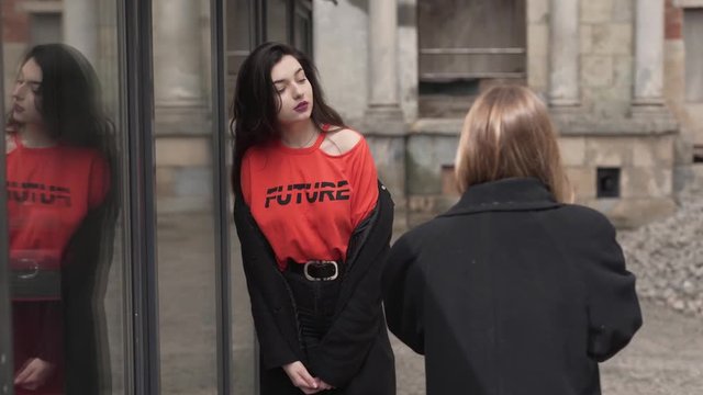 Caucasian Teenage Girl With Long Dark Hair in Red Shirt With Future Sign and Black Coat Posing By Glass Cafe Wall While Her Friend Taking Pictures With Digital Camera. Light Camera Movement