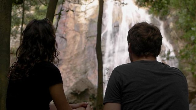 With their backs to the camera, a young couple sit and admire a waterfall, surrounded by rocks and trees