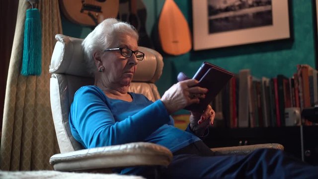 Low side view of elderly woman in comfortable chair reading an e-book.