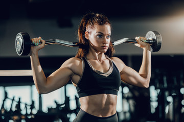 Attractive Caucasian female bodybuilder with ponytail lifting barbell while standing in gym. Train...
