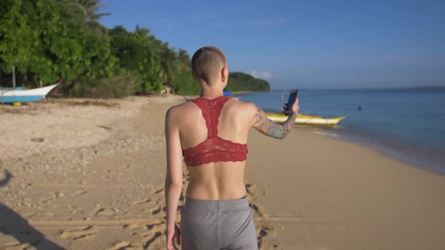 Woman walking on a tropical island beach near small boats takes a selfie and another picture of the scenery on her phone. Girl with colorful arm tattoos and shaved head walks beside the ocean. Slow mo