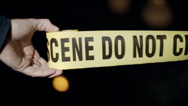 Close-up of a police officer's hand as he rolls out crime scene tape. Flashing emergency lights can be seen in the background.