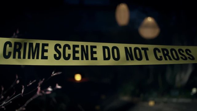 Close-up of crime scene tape as the camera tracks to the right. Lights and evidence markers can be seen in the background.