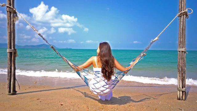 Back of brunette hair woman in white dress swinging in hammock on beach sand near turquoise sea at daytime on blue sky