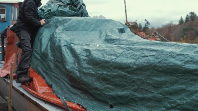 Young man unties waterproof cover of wooden boat before starting restoration maintenance