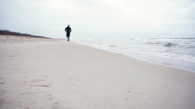 young man is running in the beatch with black tracksuit. he tries to avoid waves.