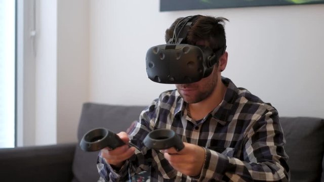 A medium shot of a young white male using Virtual Reality with controllers at home on a couch