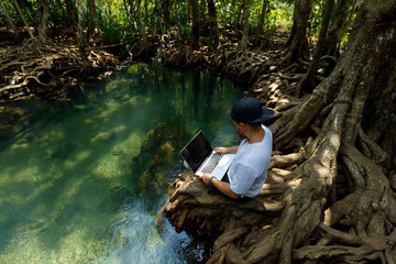 A young blogger wearing a cap and a beard is working on a holiday in a mangrove forest near the river.