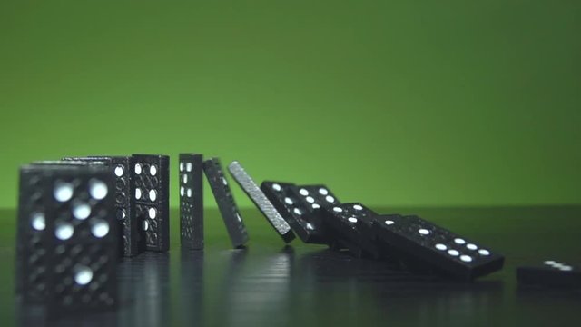 Slow motion shot of a finger nudge some black dominoes which stands in a half circle line for a chain reaction in front of a green background with plenty copy space for text.