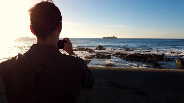 Young male on coastline documenting cargo ship sailing across the horizon. Taking photos or video with camera on sunny day.