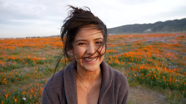 Close up on the face of a cute young woman opening her eyes and smiling with happiness in a field or meadow of wild flowers with wind blowing her hair in slow motion.