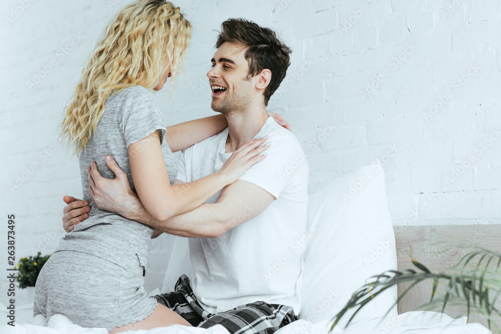 Wall mural happy handsome man looking at blonde girlfriend sitting on bed