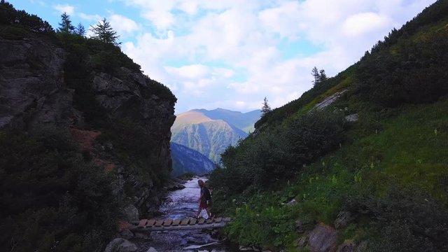 Aerial approach along a river in the mountains of Austria, passing a traveler as he crosses a bridge, and continuing over the edge of a waterfall, before revealing a spectacular vista