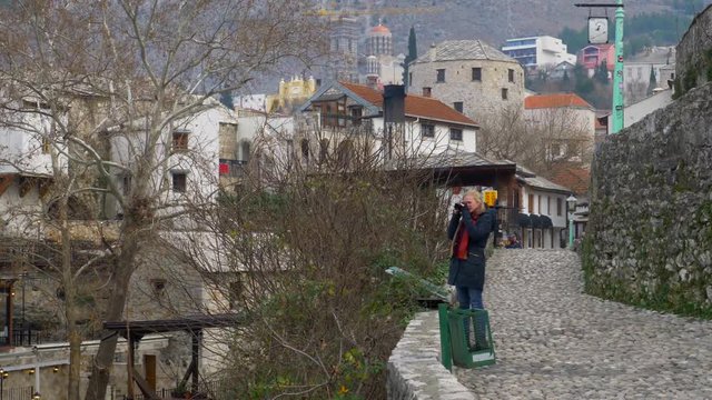 Woman in a blue coat taking a picture of barren shrubs and tree in Mostar while background shows Old TOwn Mostar