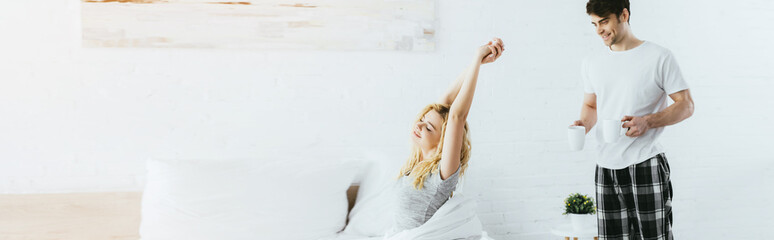panoramic shot of cheerful man holding cups near blonde woman stretching in bed