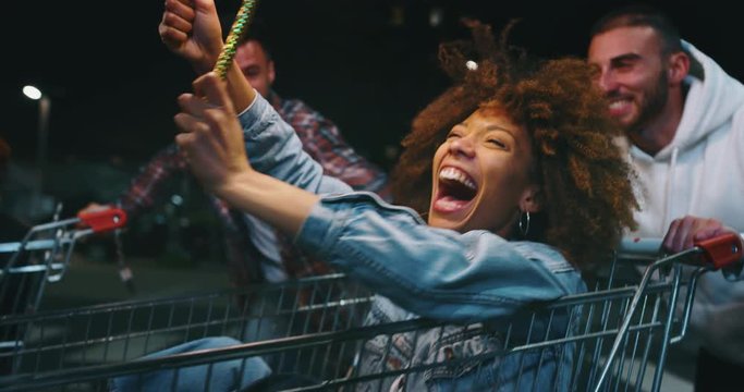 Slow motion of group of young friends of different ethnicities are having fun together racing on shopping carts and sparklers at supermarket parking at night.