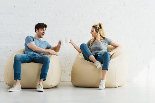 attractive blonde girl and cheerful man holding cups while sitting on bean bag chairs