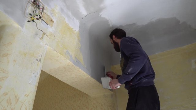 Young Caucasian Brown Haired Beard Man in Dark Purple Pullover and Track Pants is Distributing the Spackle Compound on the Ceiling Wall above the Door with a Putty Knife Spatula