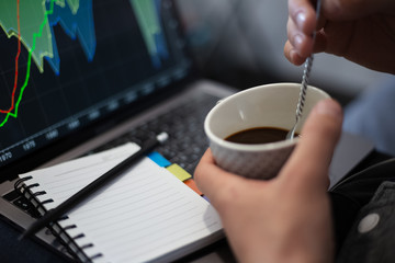 Close-up of businessman hands stirring coffee with spoon, over background of analysis finance statistic on laptop, with notepad and pencil on keyboard.