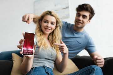 selective focus of cheerful man and blonde woman pointing with fingers at smartphone with trading courses app on screen