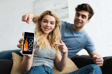 Fototapeta premium selective focus of cheerful man and blonde woman pointing with fingers at smartphone with charts and graphs on screen