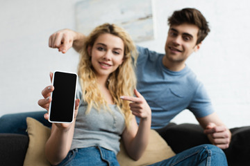 selective focus of cheerful man and blonde woman pointing with fingers at smartphone with blank screen