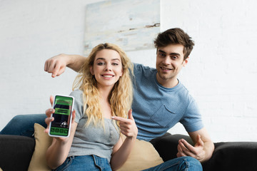 selective focus of happy man and blonde woman pointing with fingers at smartphone with booking app on screen