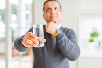Middle age man drinking glass of water at home serious face thinking about question, very confused idea