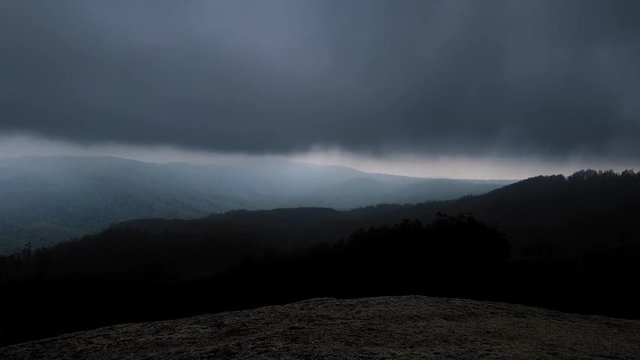 A stationary time-lapse overlooking a mountainous valley. The camera sits on a large granite boulder at the peak of a mountain. Dramatic, dark clouds and fog descended and envelope the view.