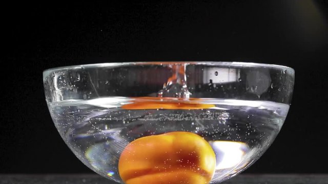 Pomodoro rosso che cade in una bacinella di vetro trasparente piena d'acqua e schizza tutto. Sfondo nero.