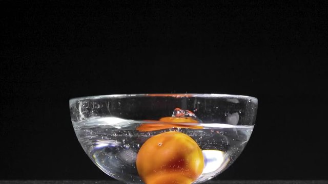 Pomodoro rosso che cade in una bacinella di vetro trasparente piena d'acqua e schizza tutto. Sfondo nero.