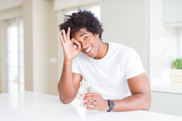 African American man drinking a glass of water at home with happy face smiling doing ok sign with hand on eye looking through fingers