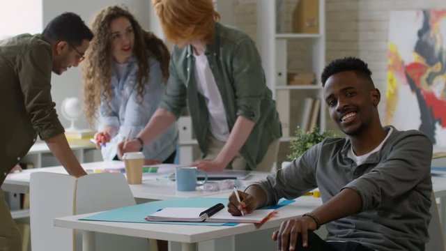 Medium panning shot of young black guy sitting at table in informal creative office and drawing sketch in notebook, with group of colleagues brainstorming on new concept in background