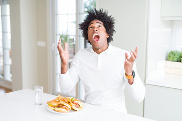 African American hungry man eating hamburger for lunch crazy and mad shouting and yelling with aggressive expression and arms raised. Frustration concept.