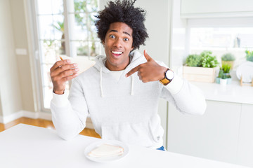 Fototapeta na wymiar African American man eating handmade sandwich at home with surprise face pointing finger to himself