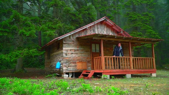 camper man watching view from balcony of wooden house while raining in forest