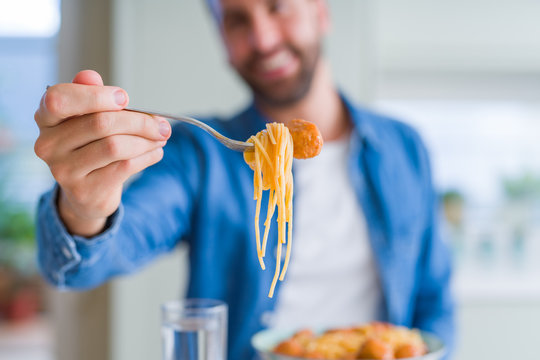 Handsome man eating pasta with meatballs and tomato sauce at home while smiling at the camera