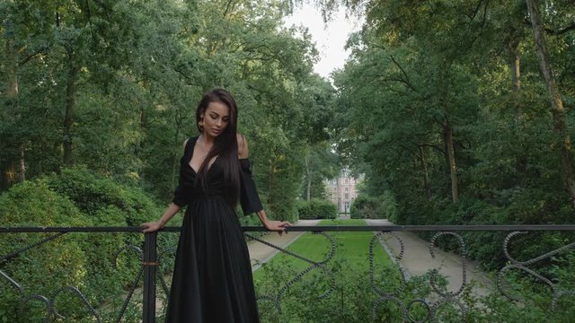 Brunette model posing in a black dress, looks to the camera and down while it's closing in, behind a beautiful castle in a national park.