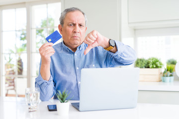 Handsome senior man shopping online using credit card and laptop at home with angry face, negative sign showing dislike with thumbs down, rejection concept