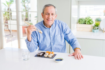 Handsome senior man eating take away sushi using chopsticks at home with a happy face standing and smiling with a confident smile showing teeth