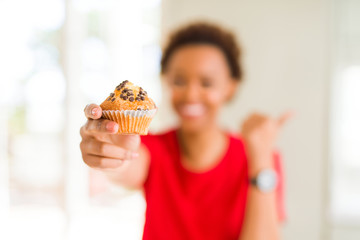 Young african american woman eating chocolate chips muffins pointing and showing with thumb up to the side with happy face smiling