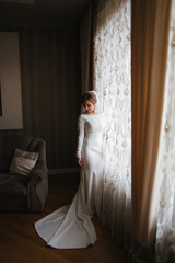 Beautiful bride standing by the window at home. Charming bride in white wedding dress