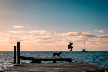 Fototapeta premium Young girl silhouette diving in the tropic ocean with her dog and a sailing boat on the background. The Bahamas 