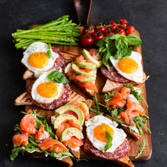 Mediterranean snacks set with brushettas, cheese variety, salami, eggs and salmon over black background on wooden board, top view