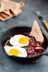 Fried eggs in a frying pan with sun dried cherry tomatoes, bacon and toasted bread for breakfast on a rustic wooden background.