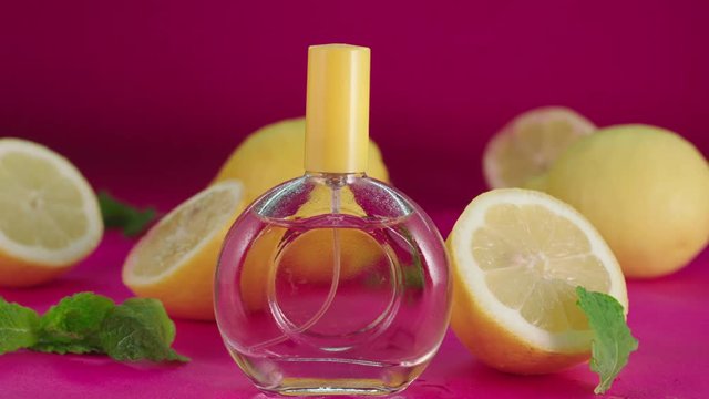 A glass bottle of perfume with an orange top, surrounded by fresh oranges and a pink background. Sprayed by a water can, cool rain effect. Real slow motion.