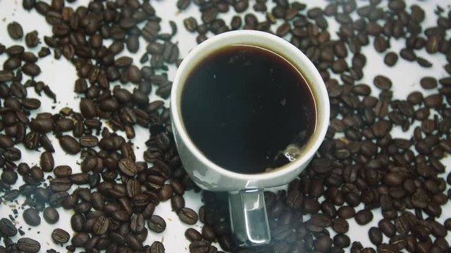 Rotating cup of coffee with steam, surrounded with brown roasted coffee beans.