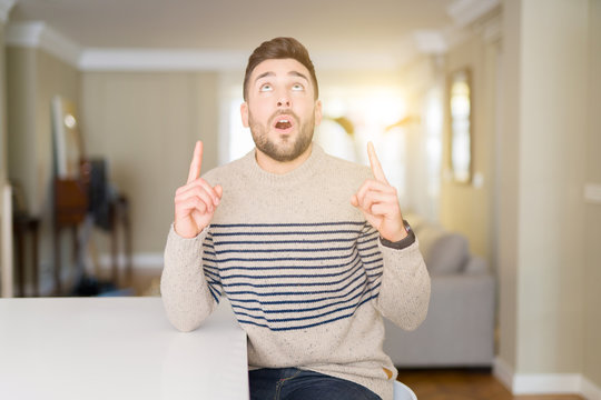Young handsome man wearing a sweater at home amazed and surprised looking up and pointing with fingers and raised arms.