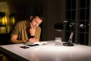 Young handsome man studying at home, reading a book at night