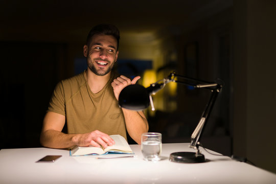 Young handsome student man studying at night at home pointing and showing with thumb up to the side with happy face smiling